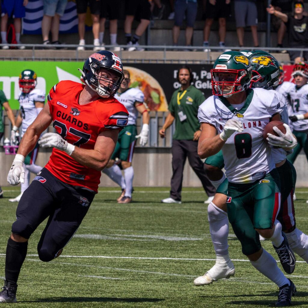 Football player runs with the ball, chased by a defender, on the field.