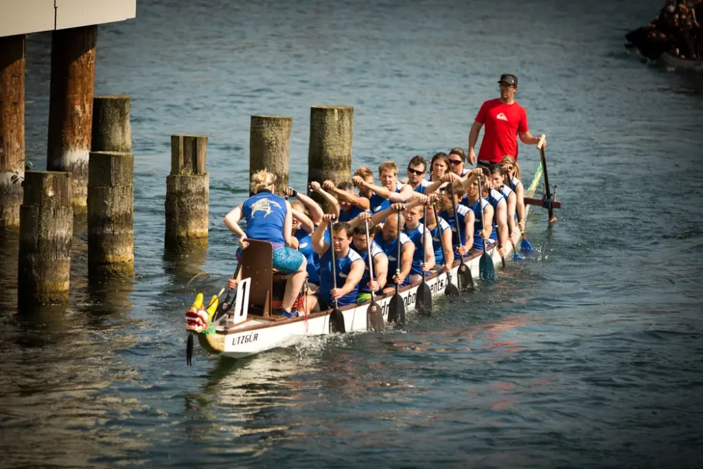 A dragon boat team paddling vigorously, led by a coach and drummer on the water.