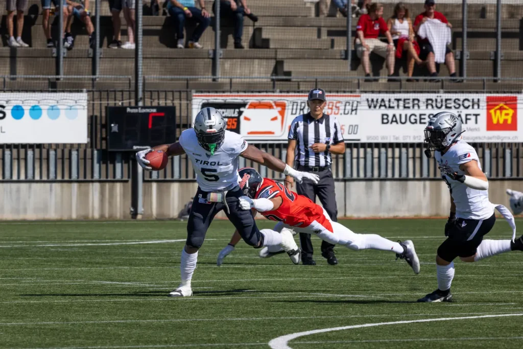 Football player with ball evades tackle; referee watches intently on sunny field.
