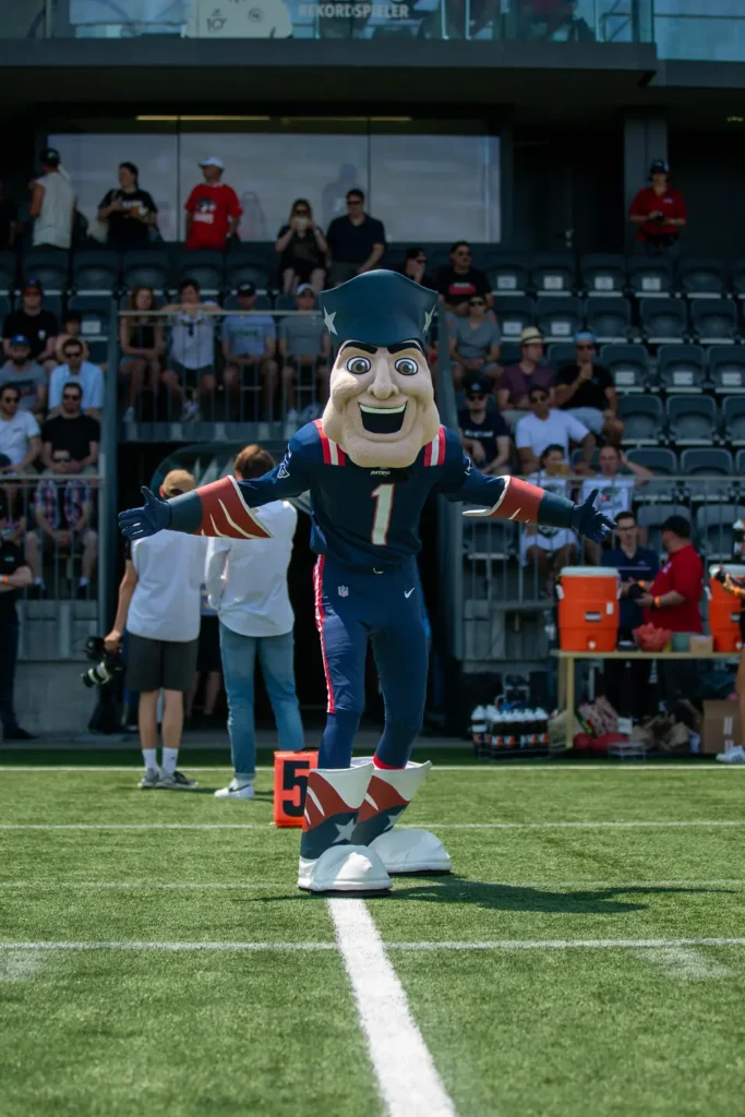 Patriots mascot, Pat Patriot, stands on a football field with outstretched arms.