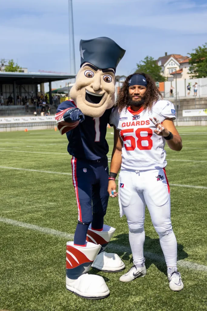 American football player with Patriots mascot on field. Bright daylight.