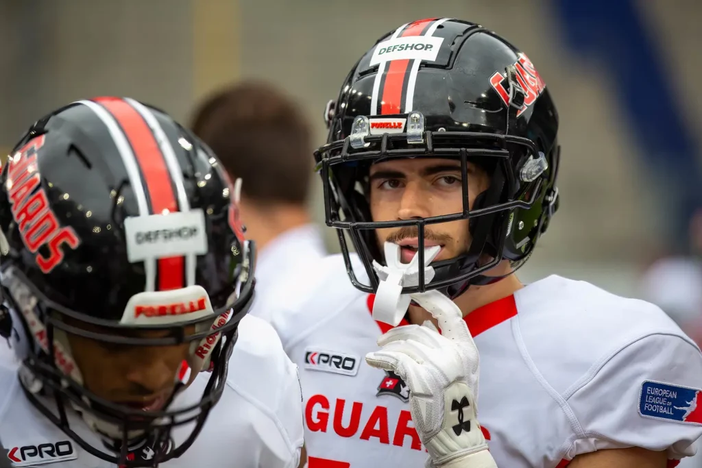 Football player in helmet and pads, holding mouthguard, looking intense.