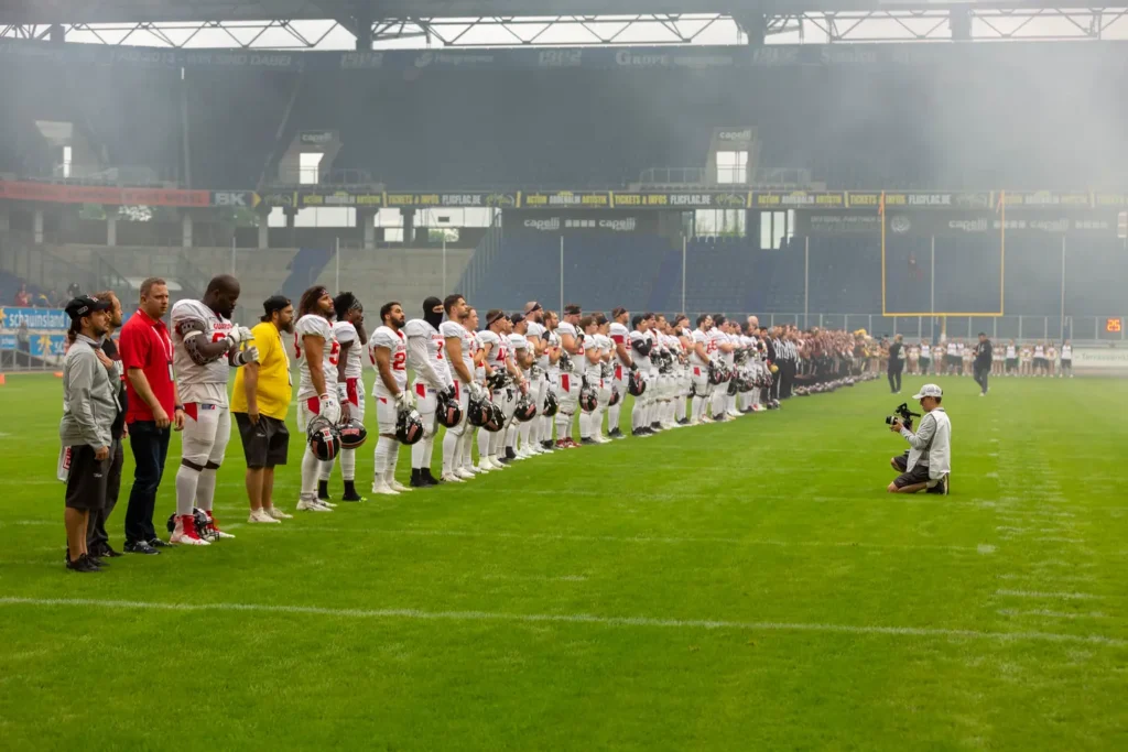 Football team stands for national anthem, photographer kneels taking picture.