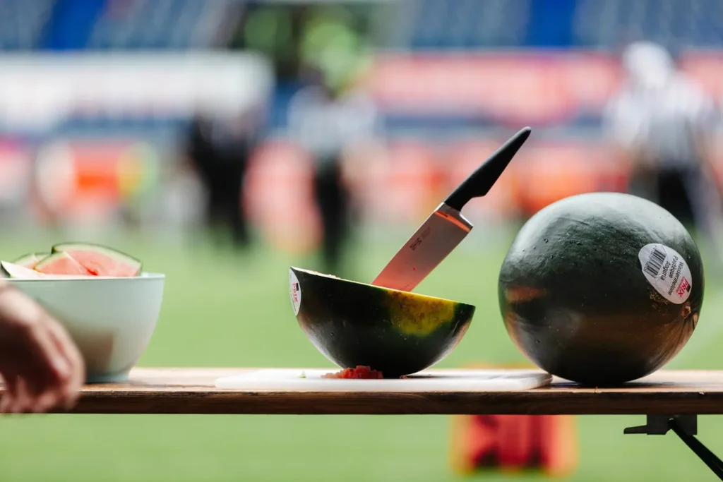 Watermelon, knife, bowl, and football field backdrop. Delicious tailgate snack.