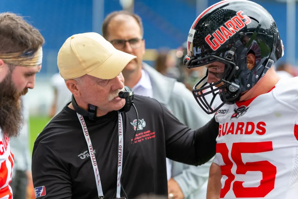 Football coach with headset talking to player in helmet on sideline.