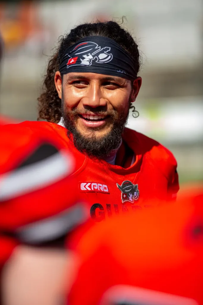 Football player with headband smiles, wearing red uniform and team logos.