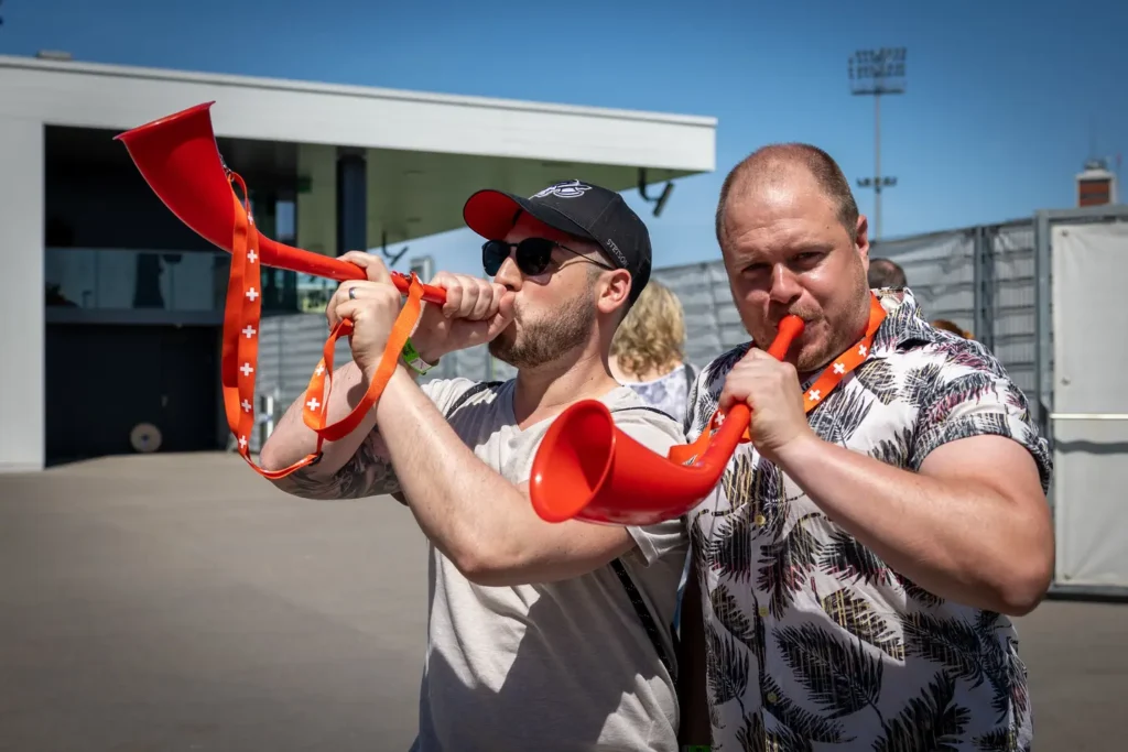 Two men blowing orange vuvuzelas with Swiss cross straps.