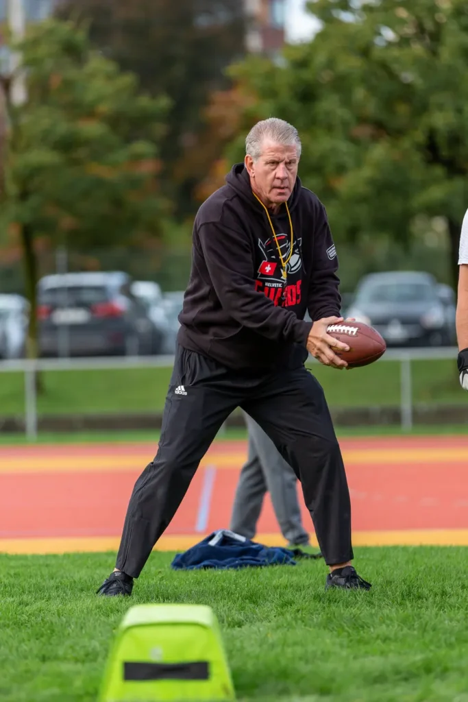 Older man in athletic wear holds a football on a green field.