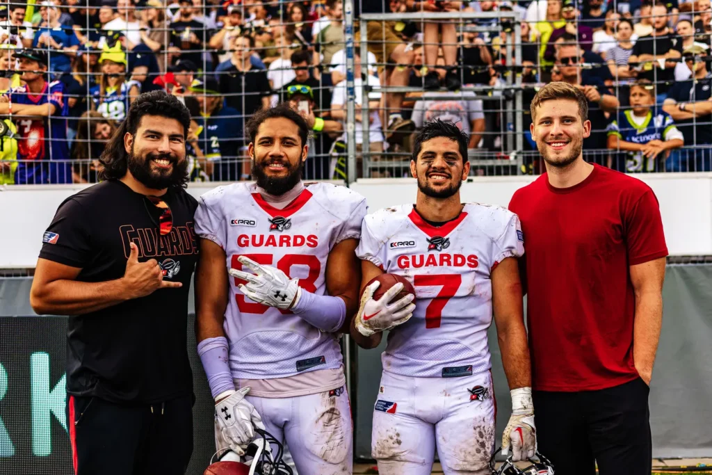 Four smiling men pose; two in American football Guards jerseys.