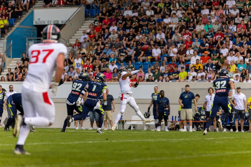 Football quarterback throwing a pass under pressure from defenders, crowd watching.