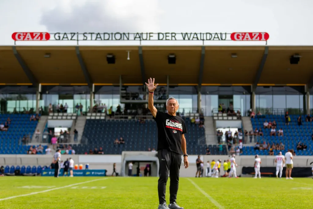 Coach Chow stands on a football field waving to the crowd.