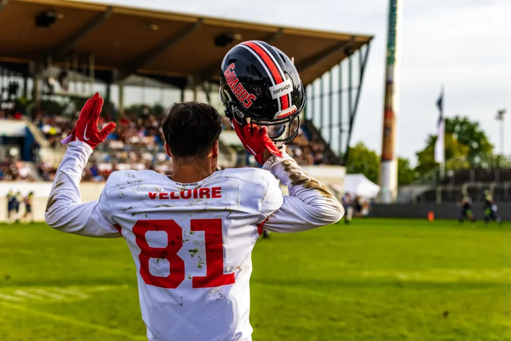 Football player in uniform waves with helmet aloft, stadium background.