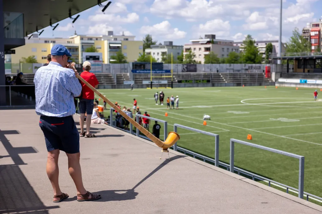 Man in cap plays alphorn at stadium overlooking football practice, outdoors.