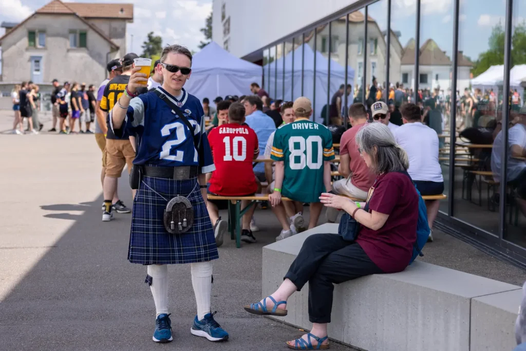 Fan wearing kilt and jersey raises beer, celebrating with other fans.