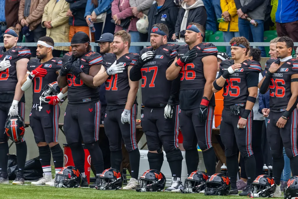 Football team in black uniforms stands with hands over hearts, helmets nearby.
