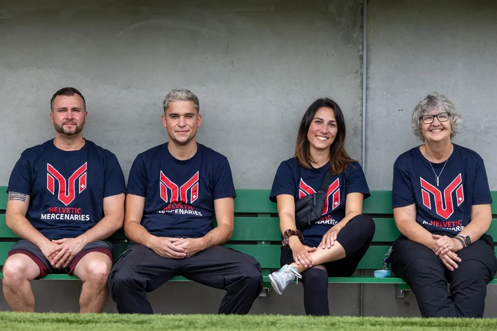 Group of four people wearing Helvetic Mercenaries shirts, sitting on bench.