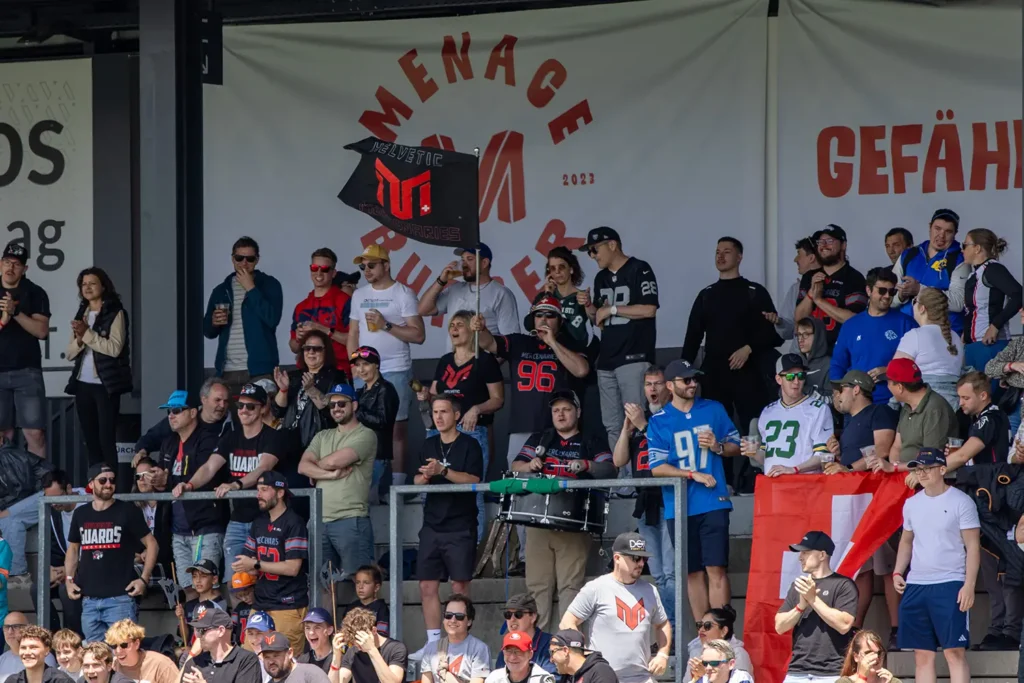 Enthusiastic fans cheer at a Swiss American football game from bleachers.