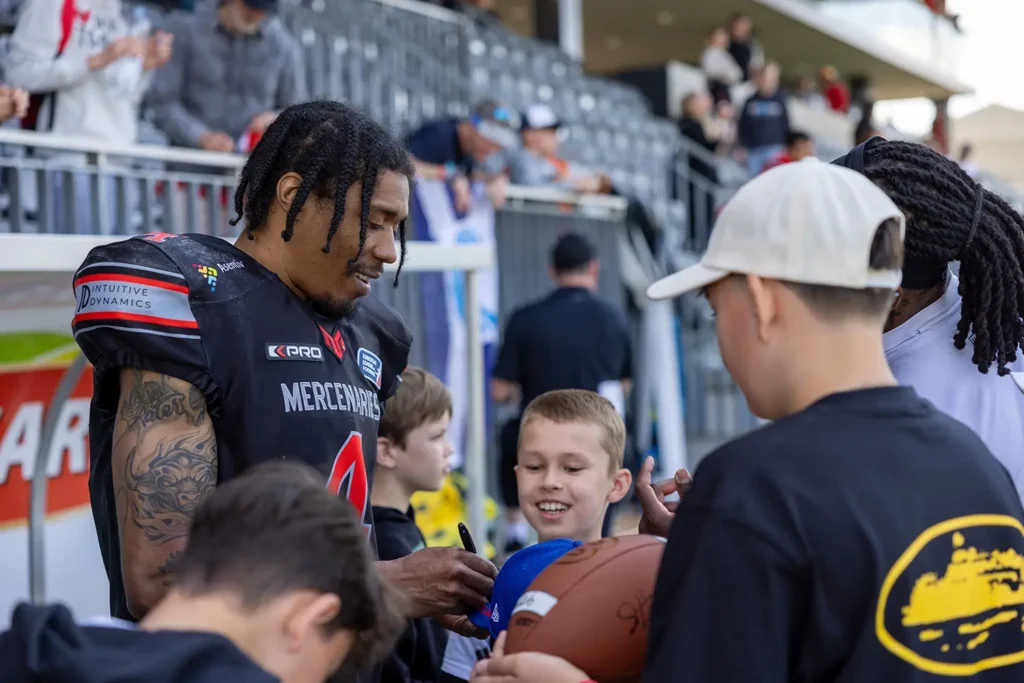 Football player in uniform signing autographs for smiling young fans at a game.