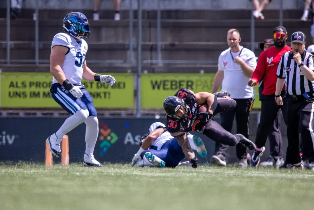 Football player tackled mid-air, helmet, pads, game officials in background.