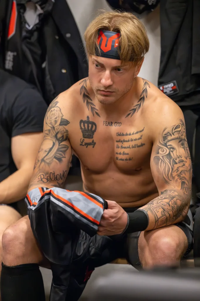 Tattooed football player with headband sitting pensively in locker room after competition.