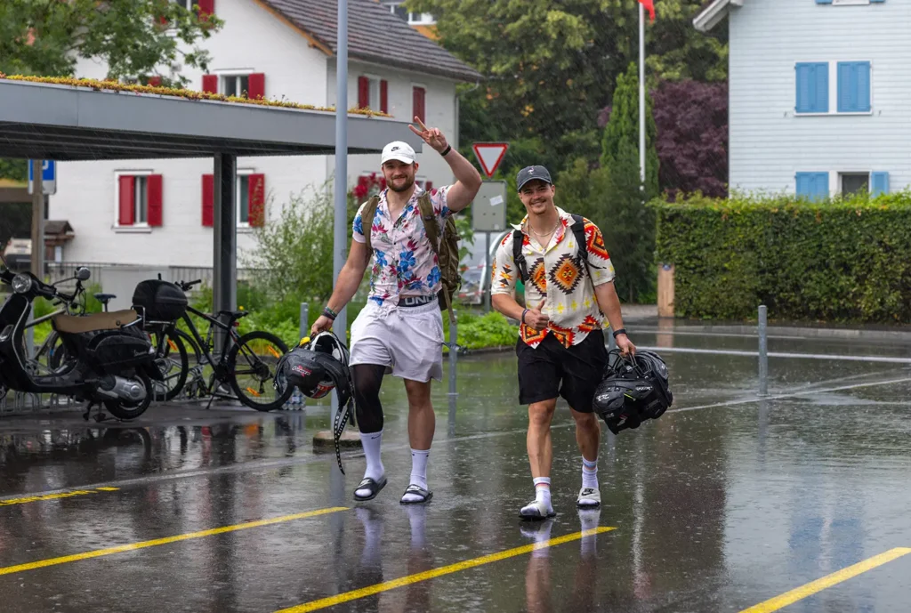 Two football players walk in rain carrying gear, summer clothes.