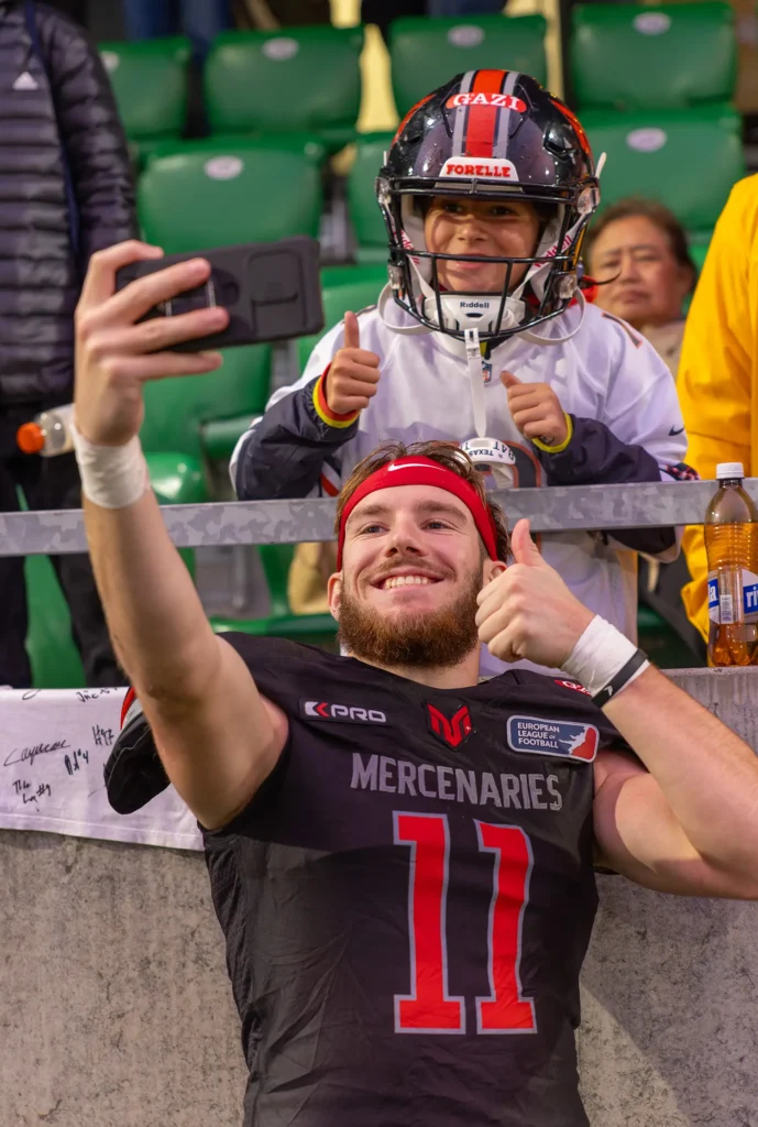 Football player taking selfie with young fan wearing helmet and jersey.