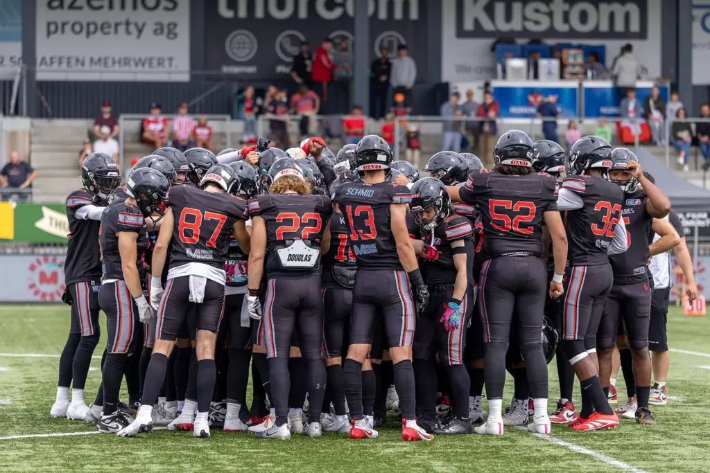 Football team in huddle; black jerseys, helmets, intense teamwork.