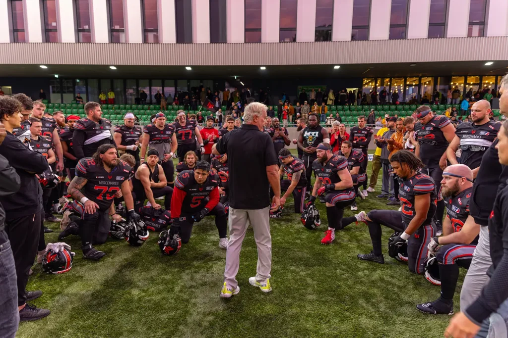 Football team gathers on field, coach facing players during team huddle.