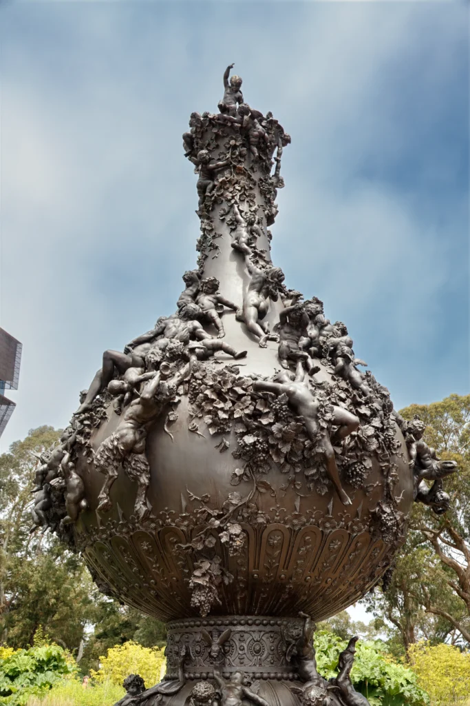 A bronze vase sculpture with figures and foliage, outdoors in a garden.