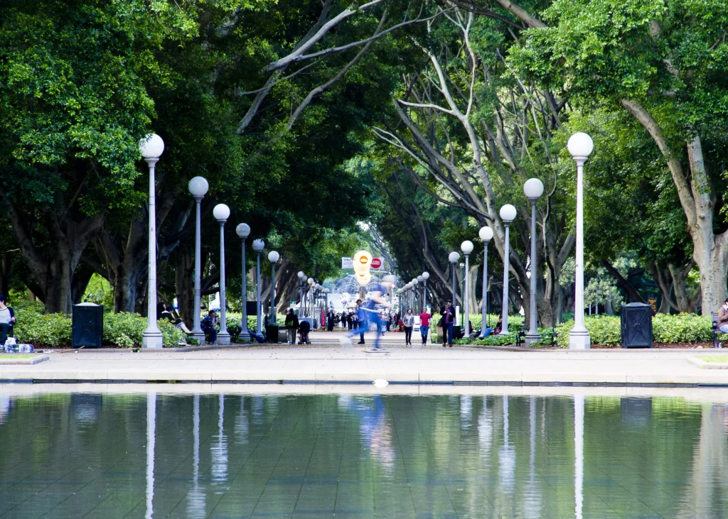 Park walkway with people, reflected in calm water, lined with trees.