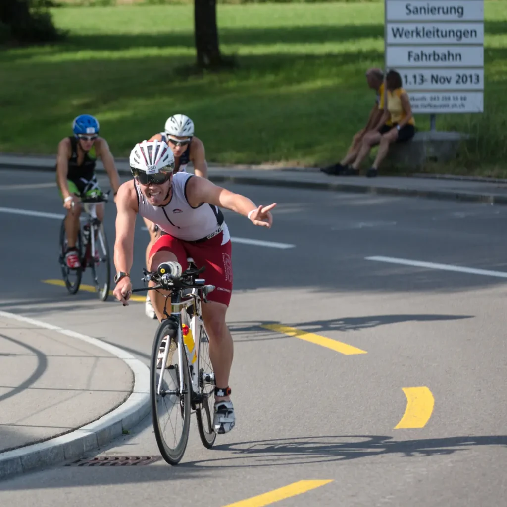 Triathlete in red and white kit gesturing while cornering on a road bike during a race