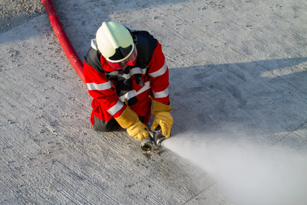 Firefighter in gear kneeling, controlling a water hose at an incident scene.