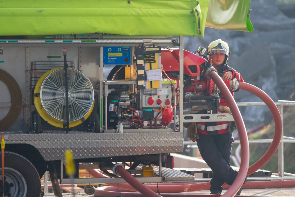 Firefighter in gear with hose near emergency vehicle.