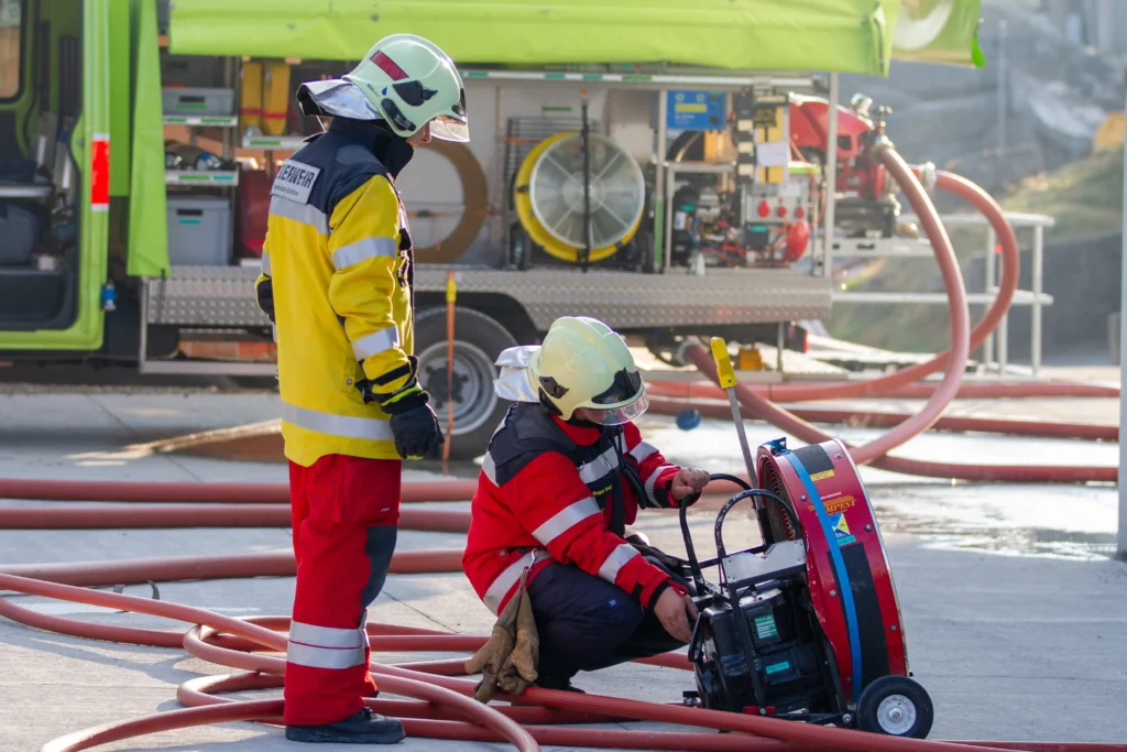 Two firefighters prepare equipment near fire truck, red hoses visible on ground.