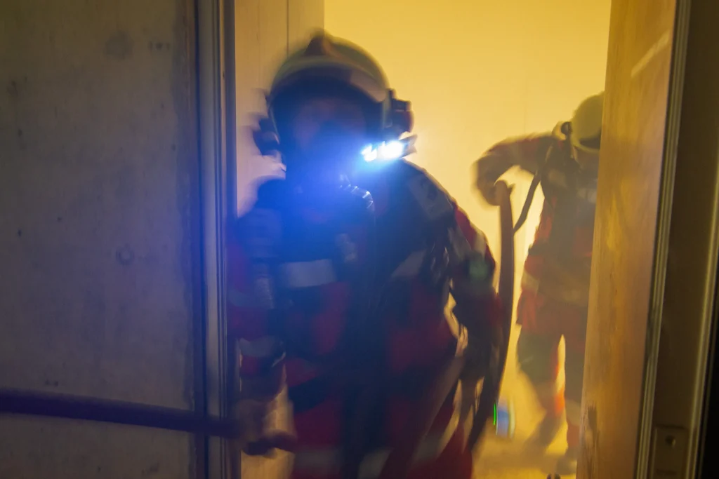 Firefighters in gear entering smoky building, lights shining, action shot.