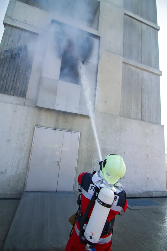 Firefighter in full gear sprays water on smoking building window.