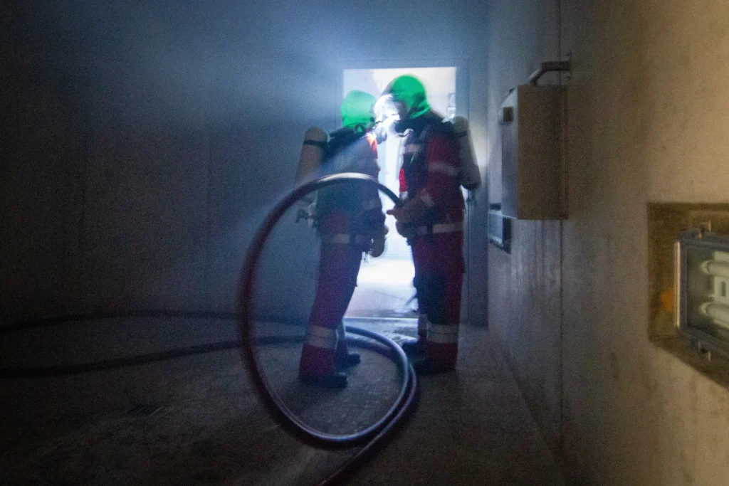Two firefighters in gear entering a smoky doorway with hose.