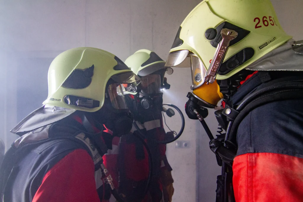 Three firefighters in full protective gear, face masks, and helmets confer closely.