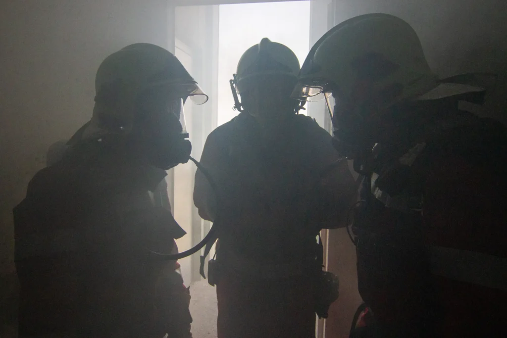 Silhouetted firefighters in gear enter smoke-filled doorway during emergency response.