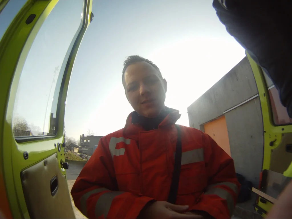 Firefighter in orange jacket stands near ambulance, looking at camera.