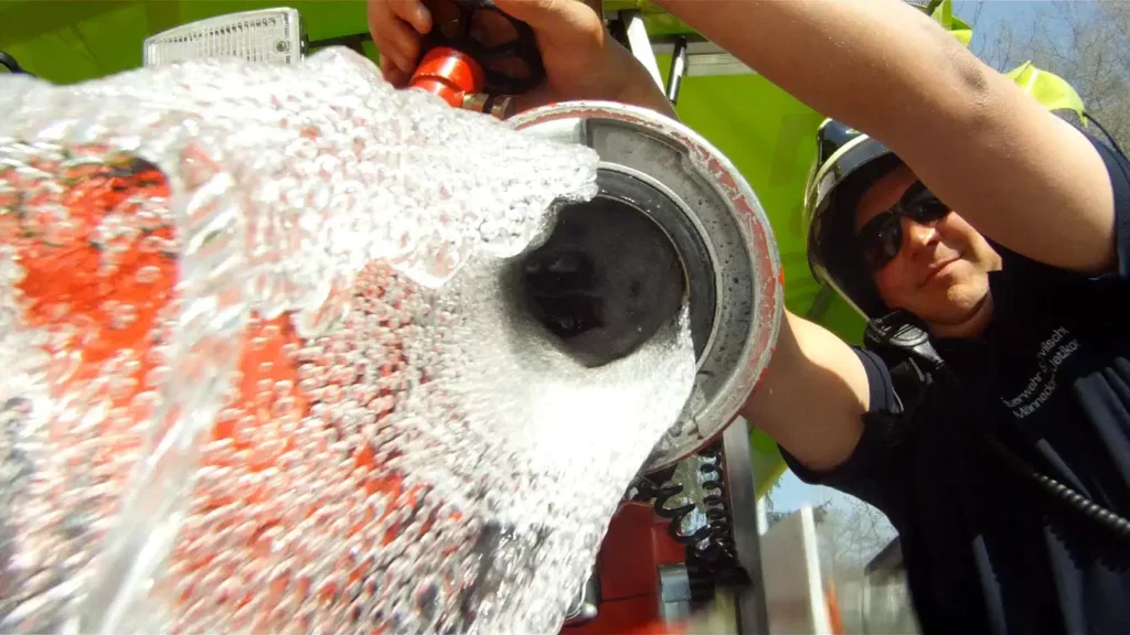 Firefighter operates a high-pressure water hose, water sprays towards the camera.