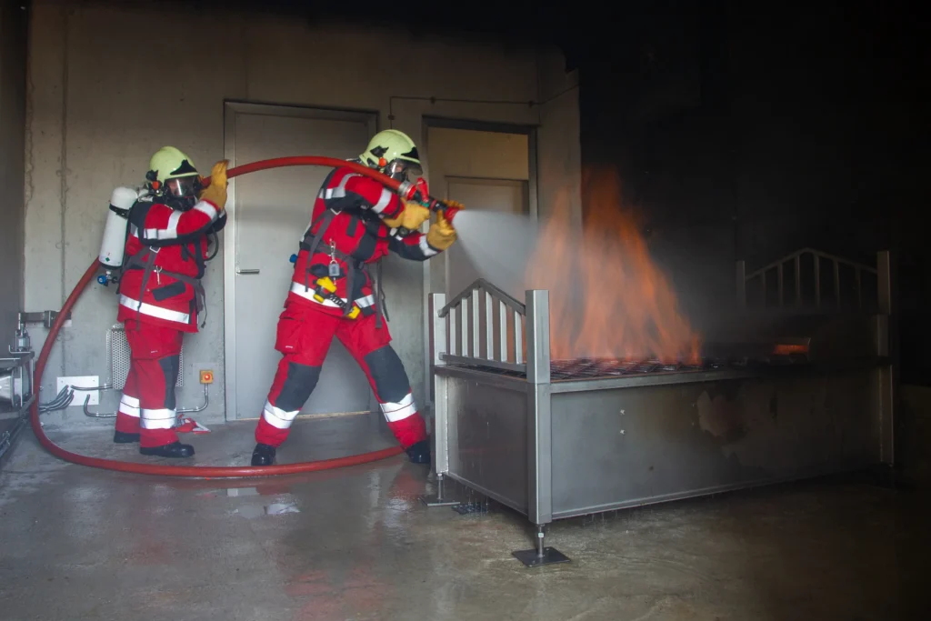 Two firefighters using hose to extinguish burning metal frame during training.