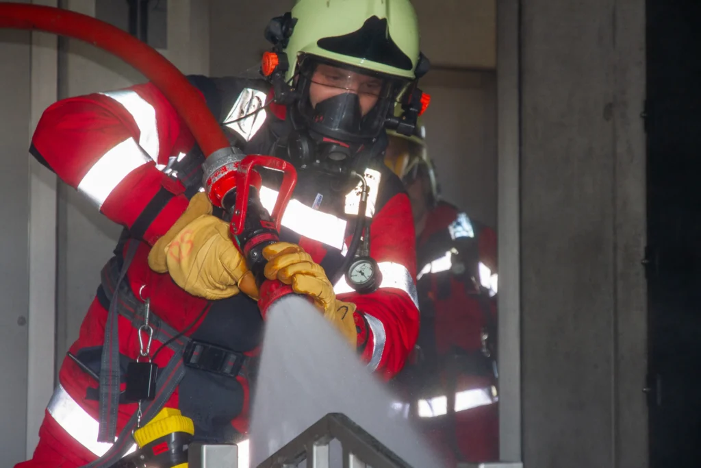 Firefighter in protective gear holding a hose, spraying water to extinguish fire.